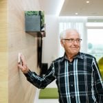Smiling wman walking into room and touch black box on the wall to demonstrate using Loxone product. NBG Digital Home. Electric Vehicle chargers, Solar and Heat Pumps, General Electrical Services in the South West.